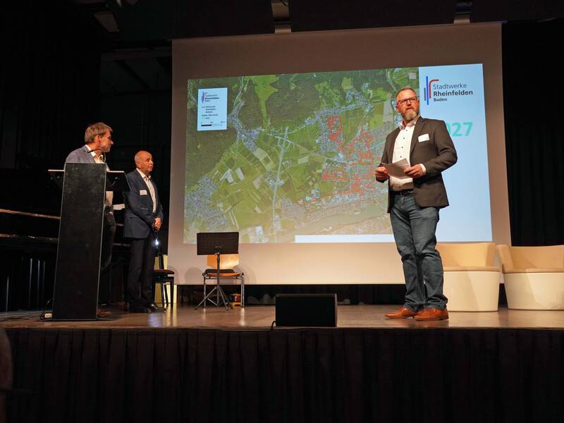Drei Männer stehen auf einer Bühne vor einer Projektion, die eine Landkarte mit der Aufschrift "Stadtwerke Rheinfelden Baden" zeigt.