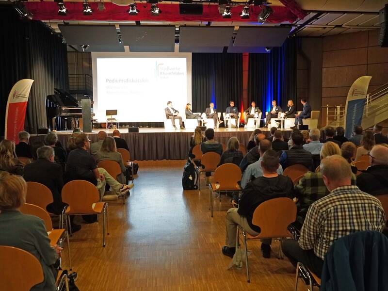 Eine Podiumsdiskussion findet in einem Veranstaltungsraum statt, in dem mehrere Personen auf der Bühne sitzen und von einem Publikum auf Stühlen beobachtet werden.