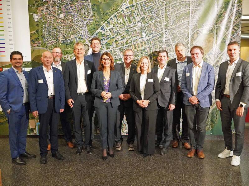 Eine Gruppe von vierzehn Menschen in formeller Kleidung steht zusammen vor einer großen Karte.