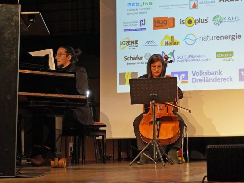 Ein Pianist und eine Cellistin spielen auf einer Bühne vor einer Leinwand mit verschiedenen Sponsorenlogos.