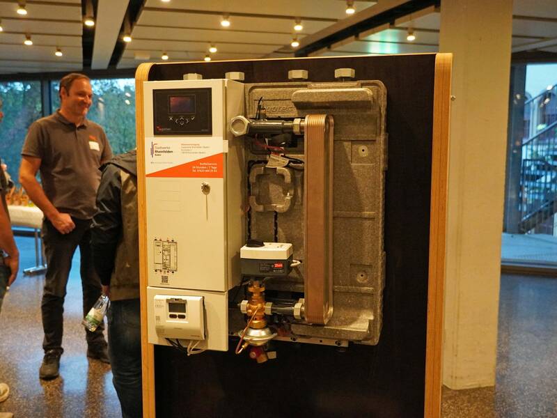 Eine industrielle Vorrichtung oder Heizsystem ist als Ausstellungsstück auf einer Messe zu sehen, während im Hintergrund mehrere Personen miteinander sprechen.