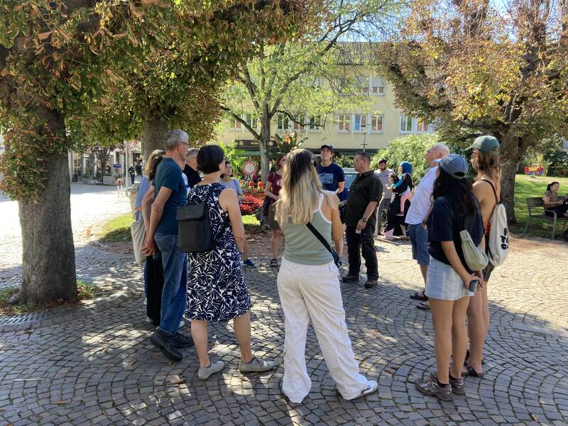 Eine Gruppe von Menschen steht in einem Kreis auf einer gepflasterten Fläche unter Bäumen und unterhält sich.