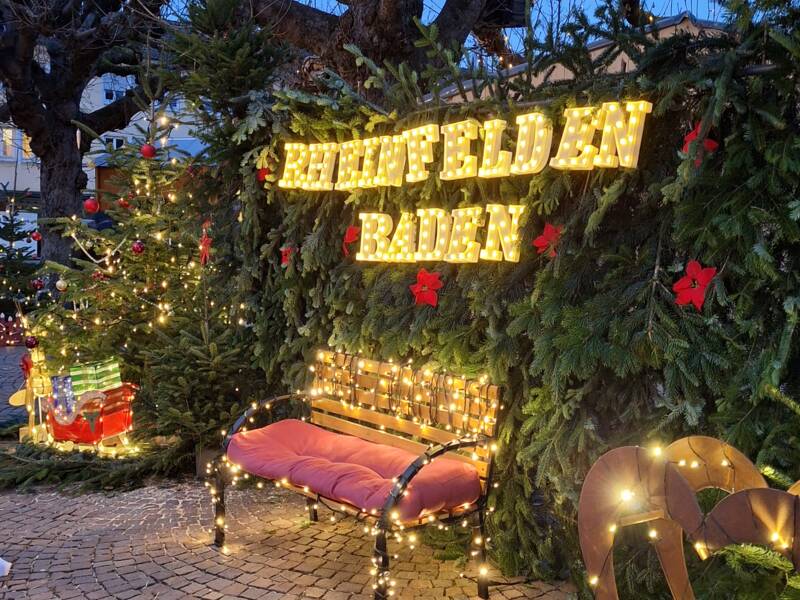 Eine festlich geschmückte Sitzbank mit Lichterketten steht vor einem mit Tannenzweigen und leuchtenden Buchstaben verzierten Hintergrund, umgeben von geschmückten Tannenbäumen.