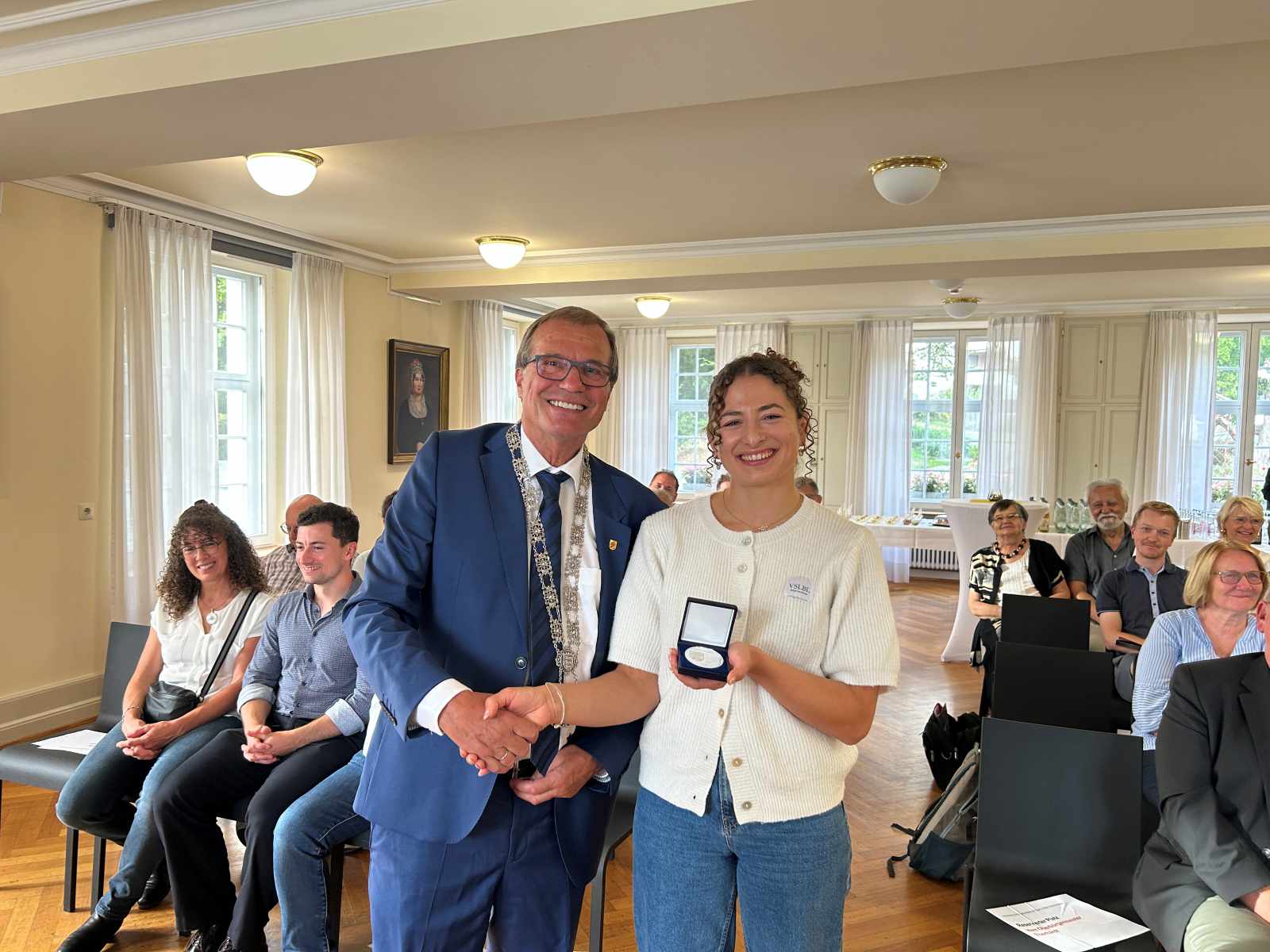 Oberbürgermeister Klaus Eberhardt gratuliert Elena Brugger zur Auszeichnung. Elena Brugger hält die Verdienstmedaille in der Hand. Beide stehen mit dem Rücken zu den Gästen. 