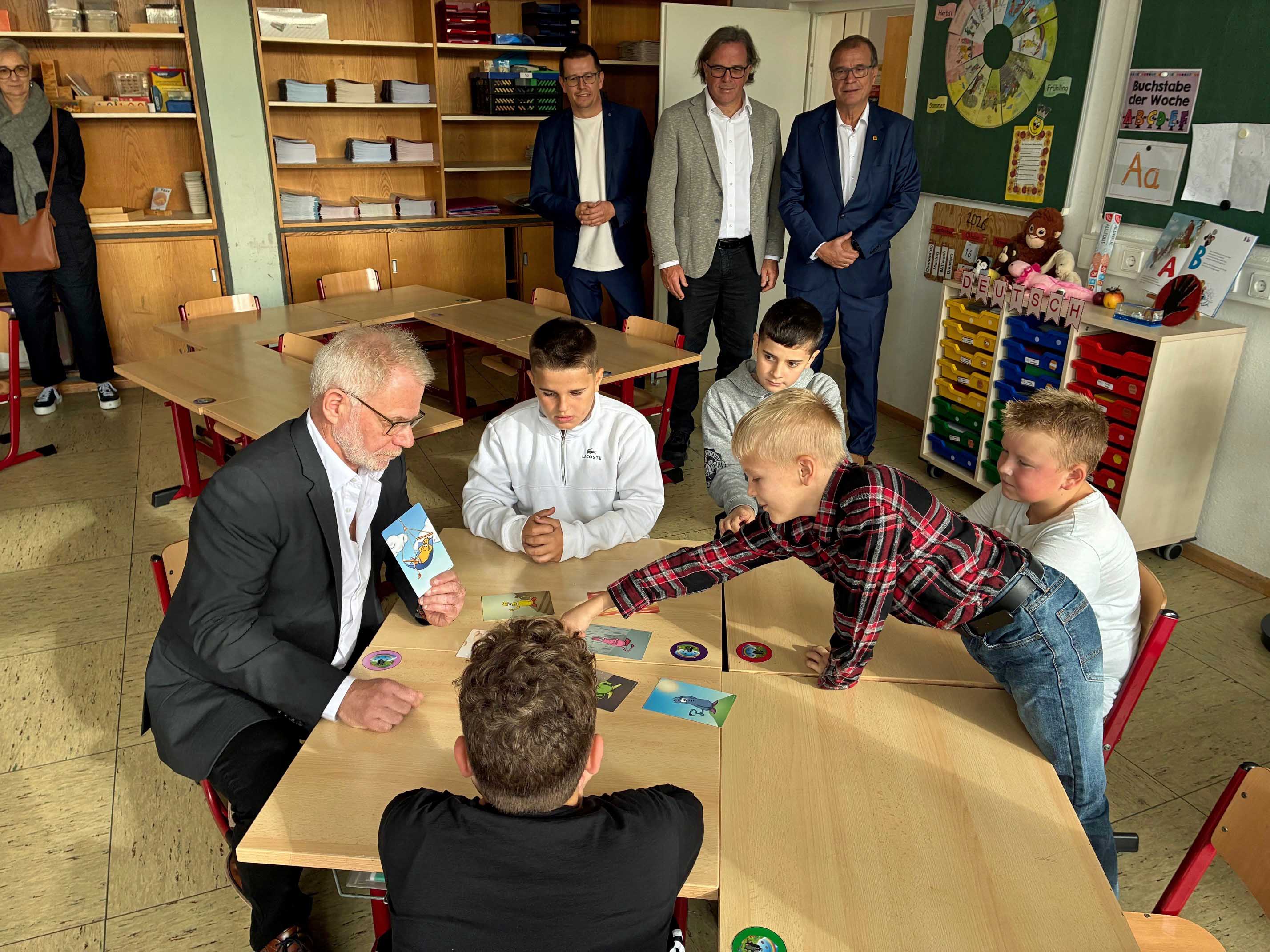 In einem Klassenzimmer sitzen und stehen mehrere Kinder mit Erwachsenen um einen Tisch herum, auf dem Karten liegen, während ein Mann in einem Anzug eine Karte hochhält.